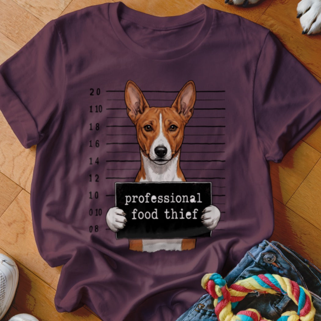 Basenji dog mugshot with "professional food thief" sign on maroon t-shirt.