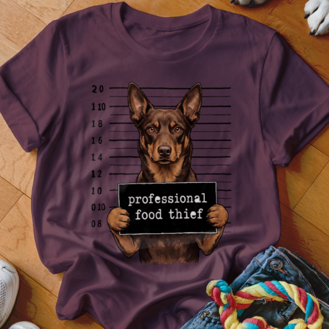 Brown dog in mugshot with "professional food thief" sign on burgundy t-shirt.