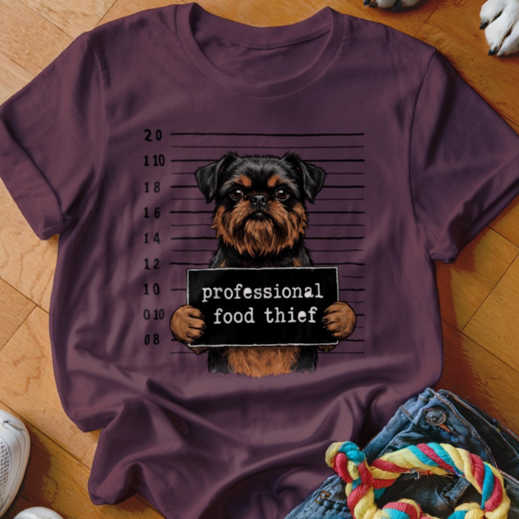 Dog with "professional food thief" sign in front of mugshot backdrop on maroon t-shirt.