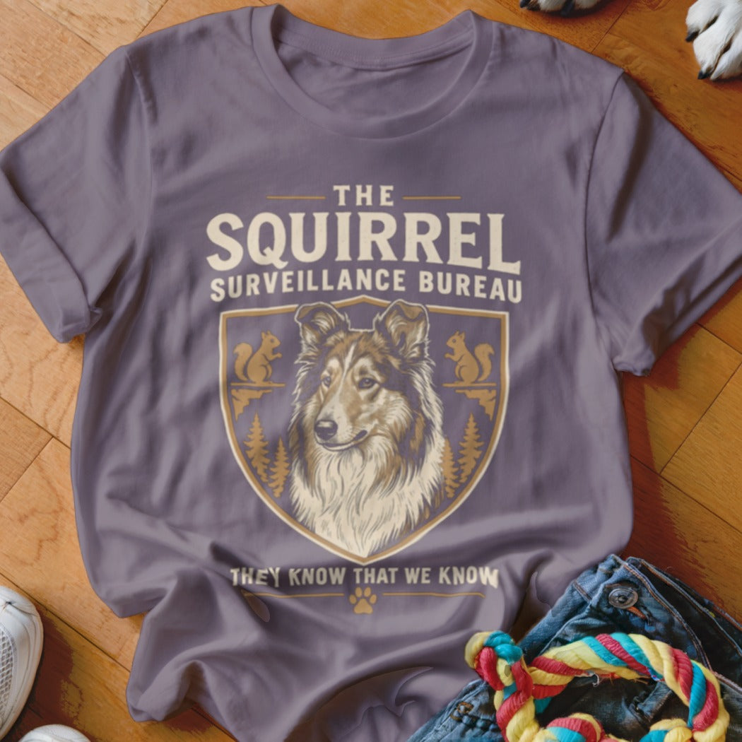 A purple t-shirt featuring a crest with a dog, squirrels, trees, and text that reads The Squirrel Surveillance Bureau.