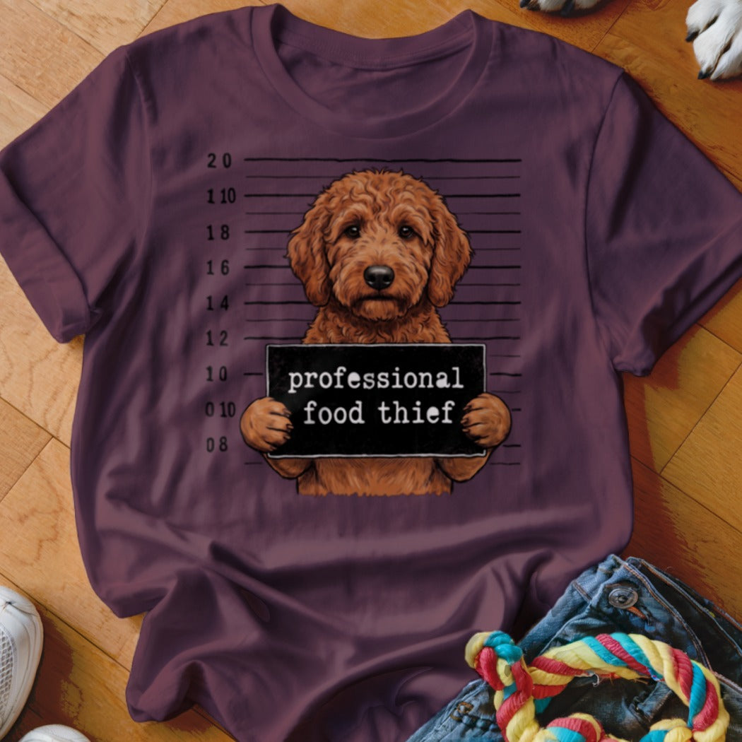 Goldendoodle mugshot with "professional food thief" sign on t-shirt.