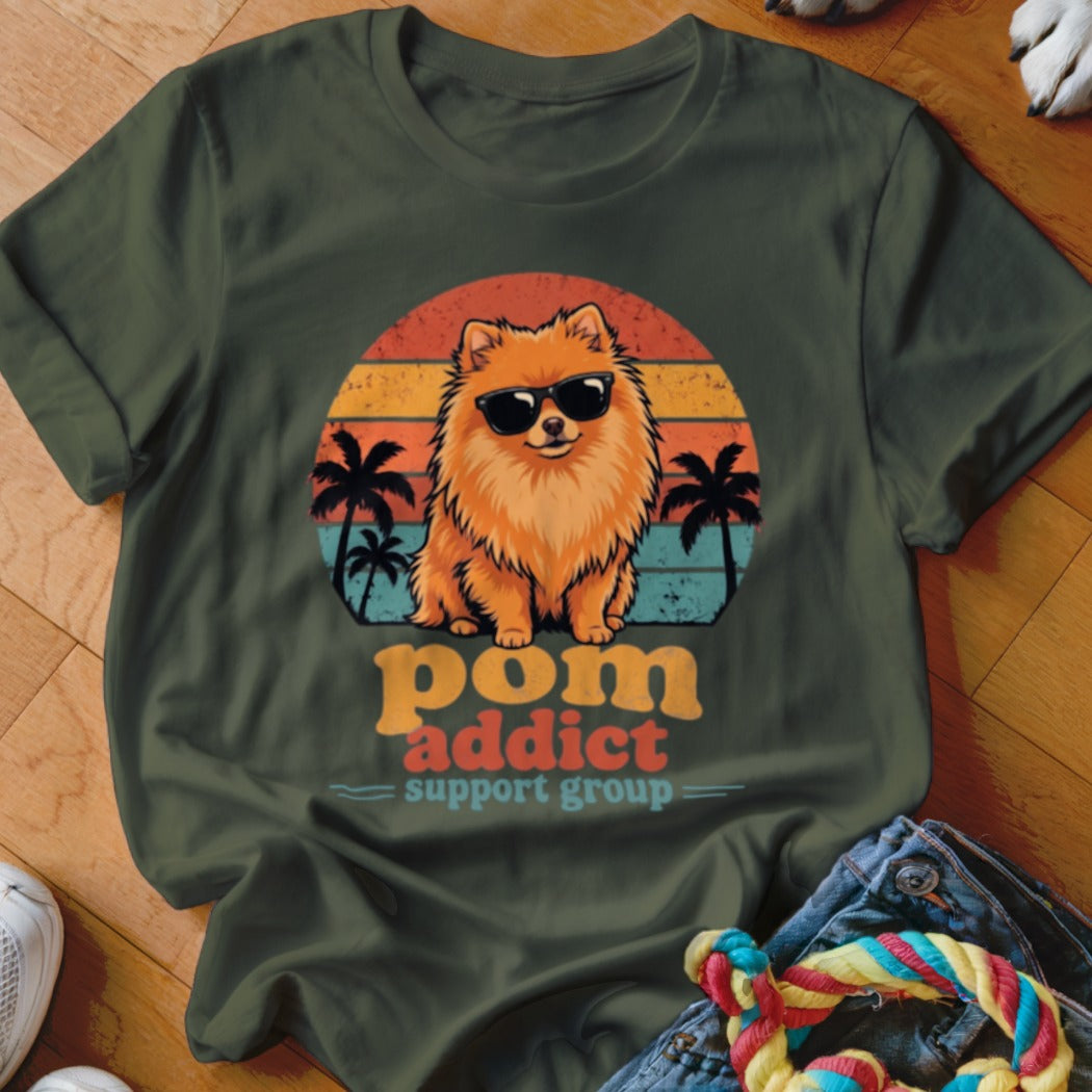 Dark green t-shirt featuring a retro graphic of a Pomeranian dog wearing sunglasses, palm trees, and sunset stripes, with the text "pom addict support group".