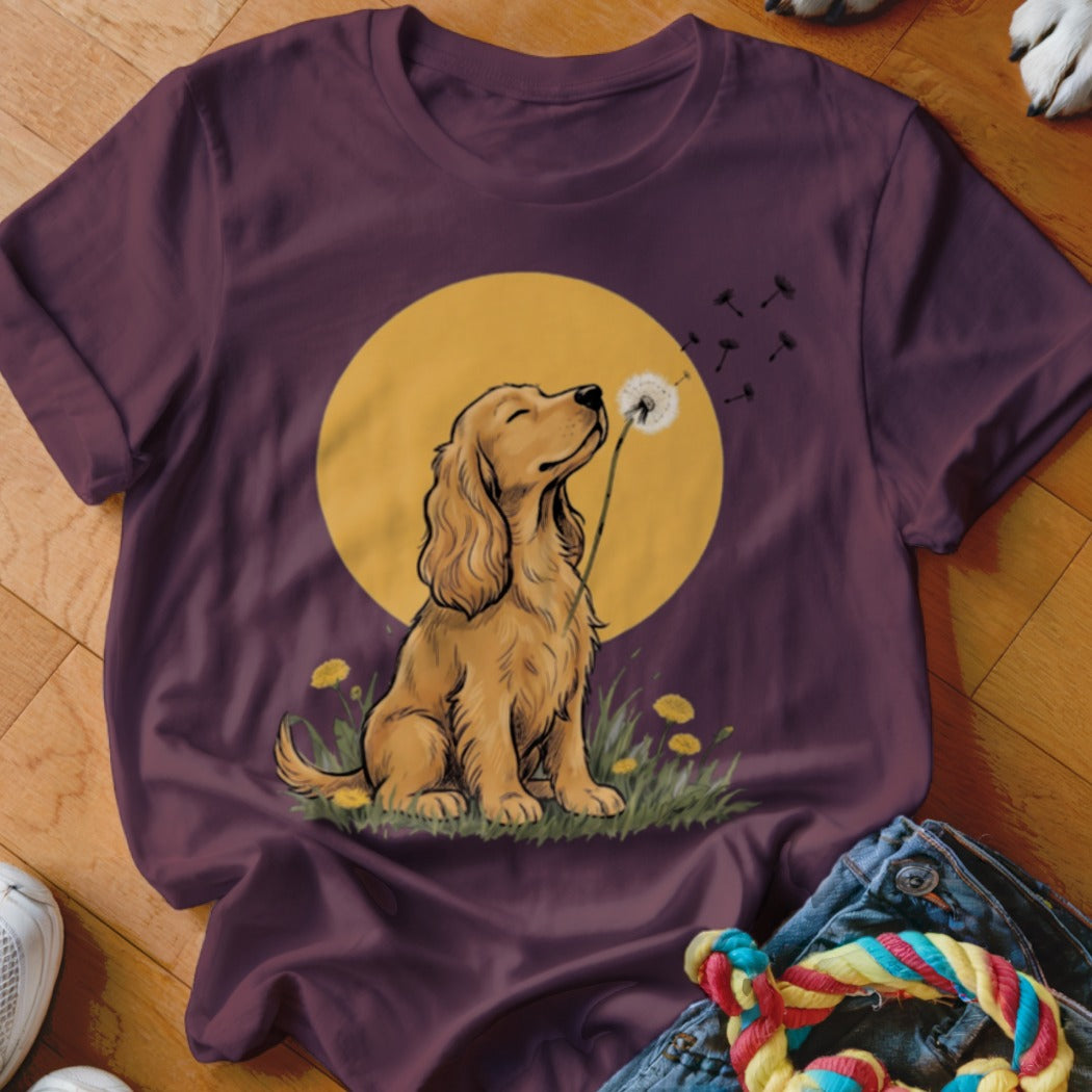 A plum-colored t-shirt features a graphic of a golden spaniel blowing a dandelion against a yellow sun.