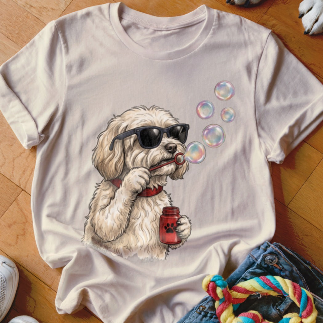 A cream-colored t-shirt featuring a graphic of a fluffy white dog wearing sunglasses and blowing bubbles.