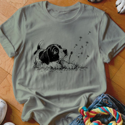 Sage green t-shirt featuring a black and white illustration of a puppy looking up at dandelion seeds.