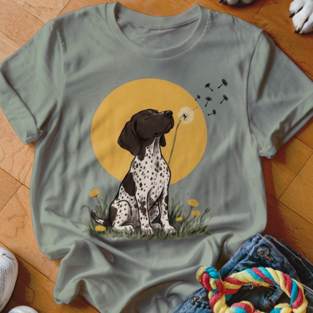 Sage green t-shirt featuring an illustration of a spotted puppy sitting in grass blowing dandelion seeds.