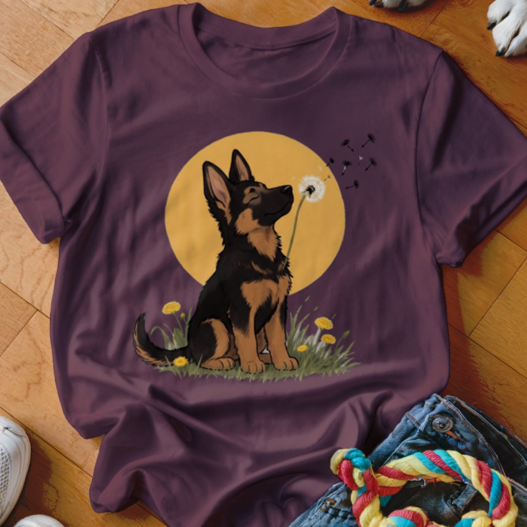 Maroon t-shirt featuring a cartoon illustration of a German Shepherd puppy blowing a dandelion against a yellow sun.