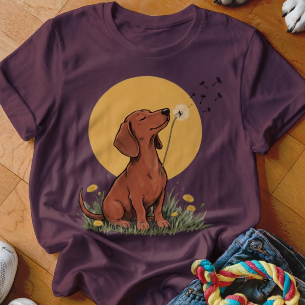 Plum-colored t-shirt featuring a graphic of a dachshund sitting in grass, blowing dandelion seeds in front of a yellow moon.