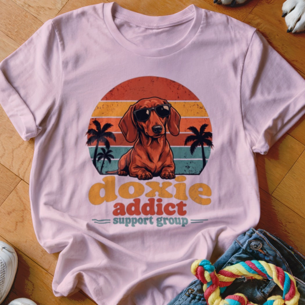 A light pink t-shirt with a retro dachshund graphic featuring sunglasses, palm trees, and text reading doxie addict support group.