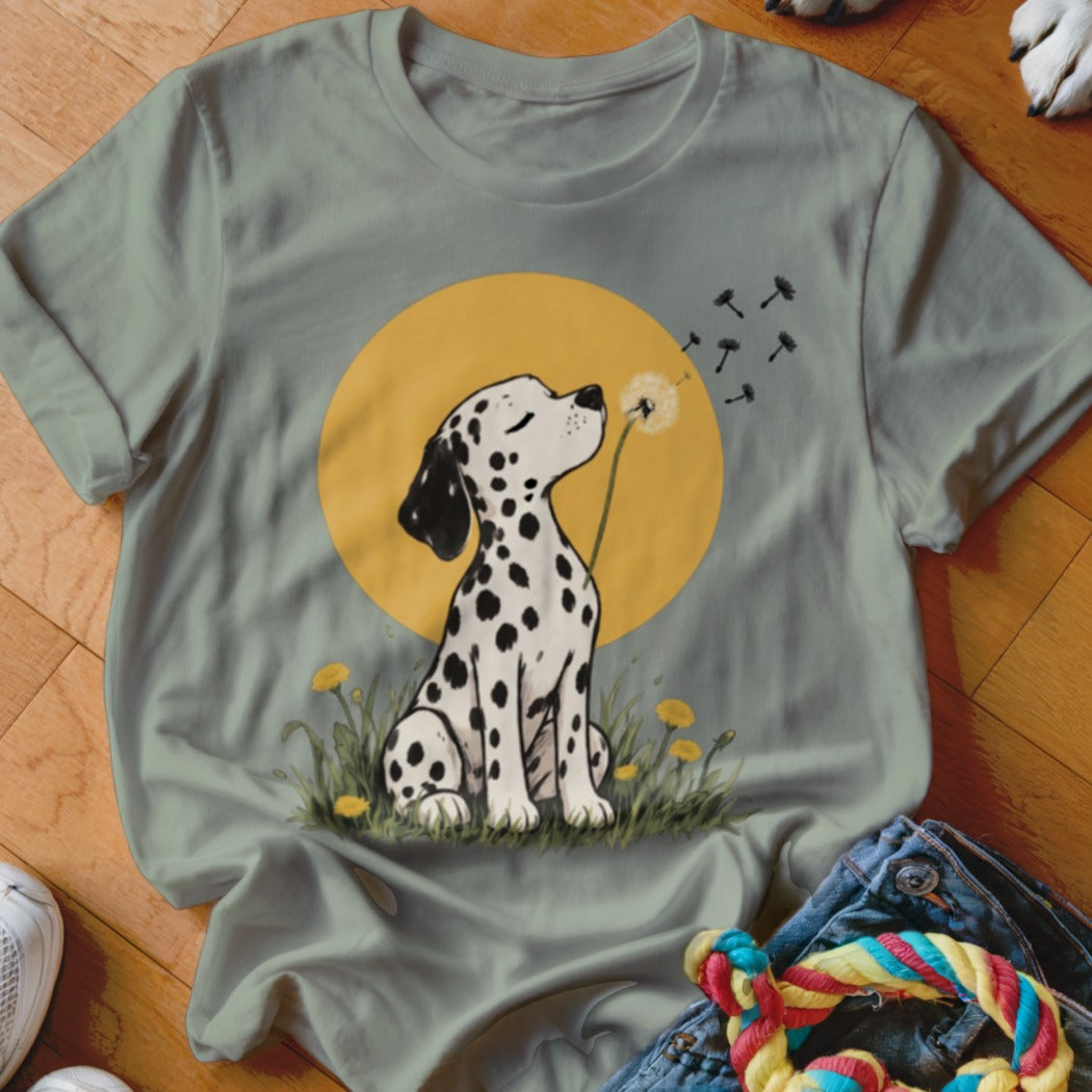 Sage green t-shirt featuring a graphic of a Dalmatian puppy blowing dandelion seeds against a yellow sun.