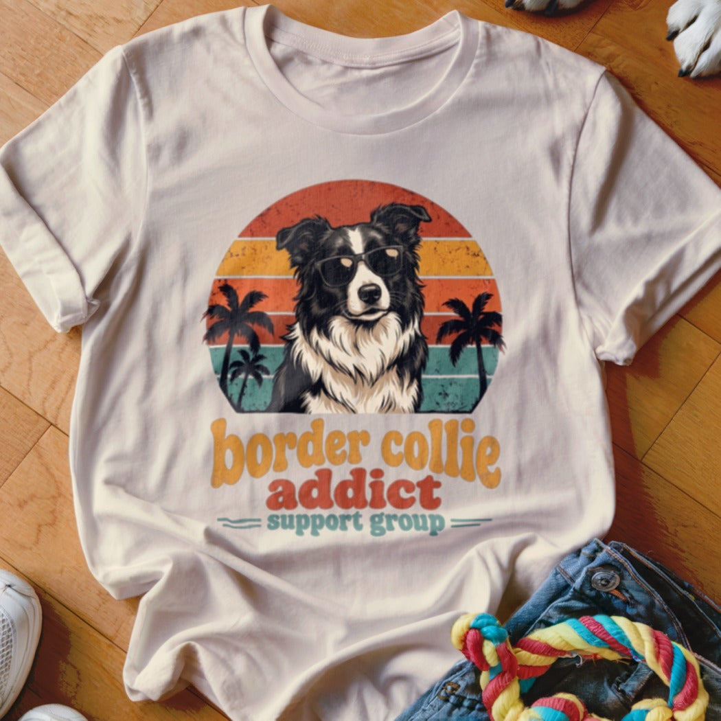 A beige t-shirt featuring a retro graphic of a sunglasses-wearing border collie, palm trees, and text reading "border collie addict support group".