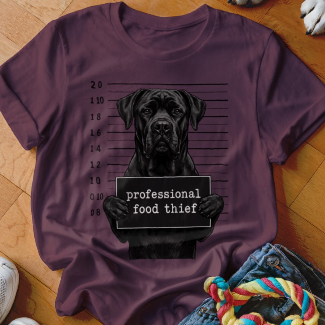 Black dog mugshot with "professional food thief" sign on a maroon t-shirt.