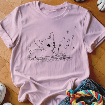 A light pink t-shirt featuring a black line art graphic of a puppy in grass blowing dandelion seeds.