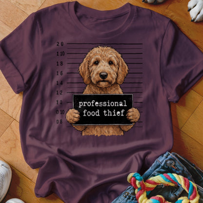 Goldendoodle mugshot with "professional food thief" sign on maroon t-shirt.