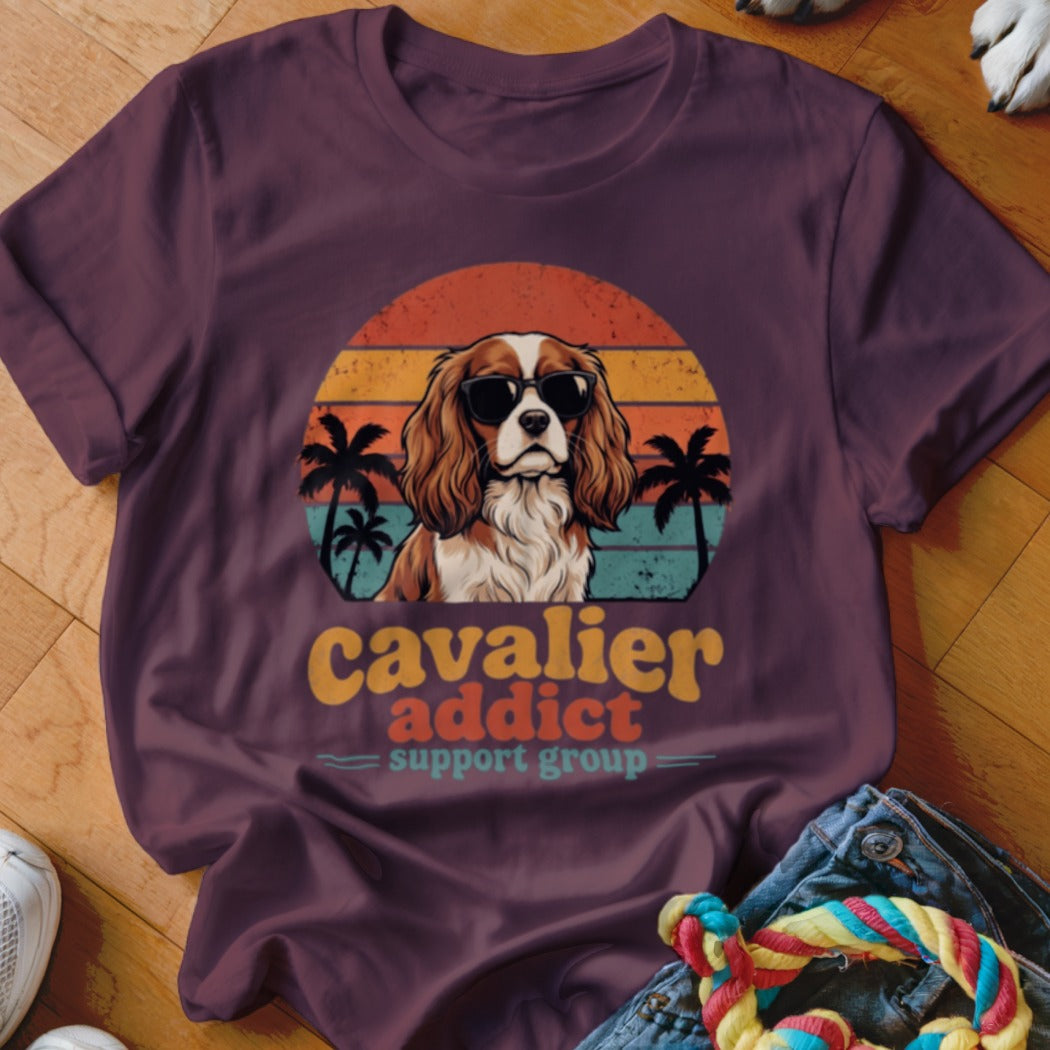 A dark purple t-shirt featuring a retro graphic of a Cavalier King Charles Spaniel wearing sunglasses against a sunset and palm trees, with the text "Cavalier Addict Support Group".