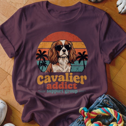 A dark purple t-shirt featuring a retro graphic of a Cavalier King Charles Spaniel wearing sunglasses against a sunset and palm trees, with the text "Cavalier Addict Support Group".