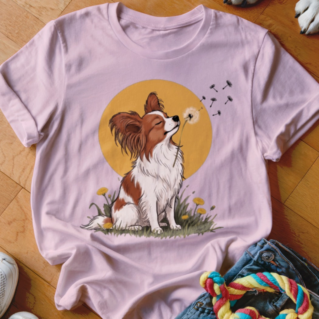 A pink t-shirt featuring an illustration of a papillon dog blowing dandelion seeds against a yellow circle.