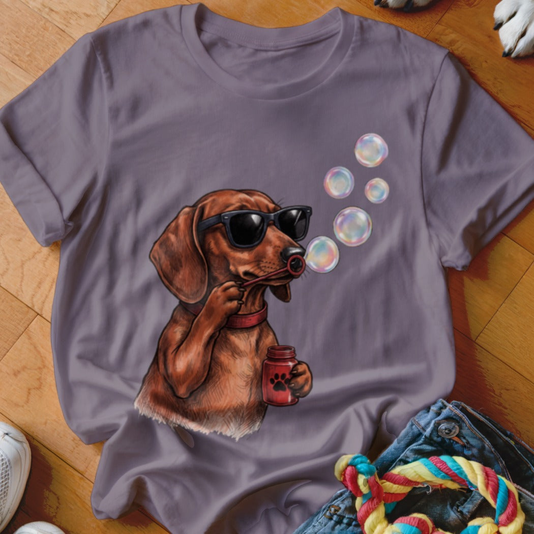 A dusty purple t-shirt features a graphic of a dachshund wearing sunglasses and blowing bubbles.