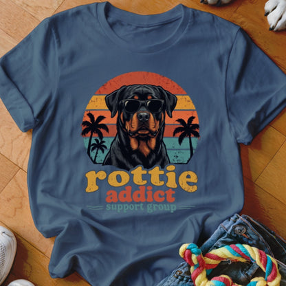 A blue t-shirt featuring a Rottweiler wearing sunglasses in front of a sunset graphic, with text that reads "Rottie Addict Support Group".