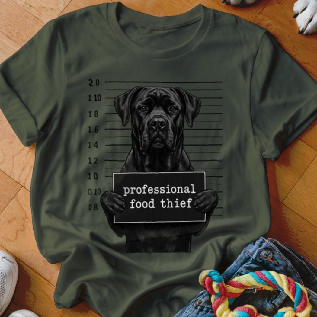 Black dog with mugshot sign reading "professional food thief" on t-shirt.