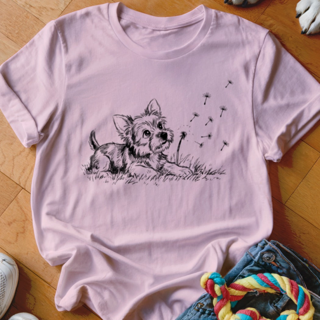 A pink t-shirt features a black graphic of a puppy in grass watching dandelion seeds blow.