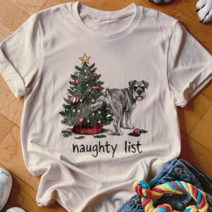 A cream-colored t-shirt featuring a graphic of a Schnauzer urinating on a Christmas tree, with the text naughty list underneath.