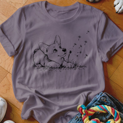 A mauve crewneck t-shirt featuring a black line drawing of a corgi puppy watching blowing dandelion seeds.