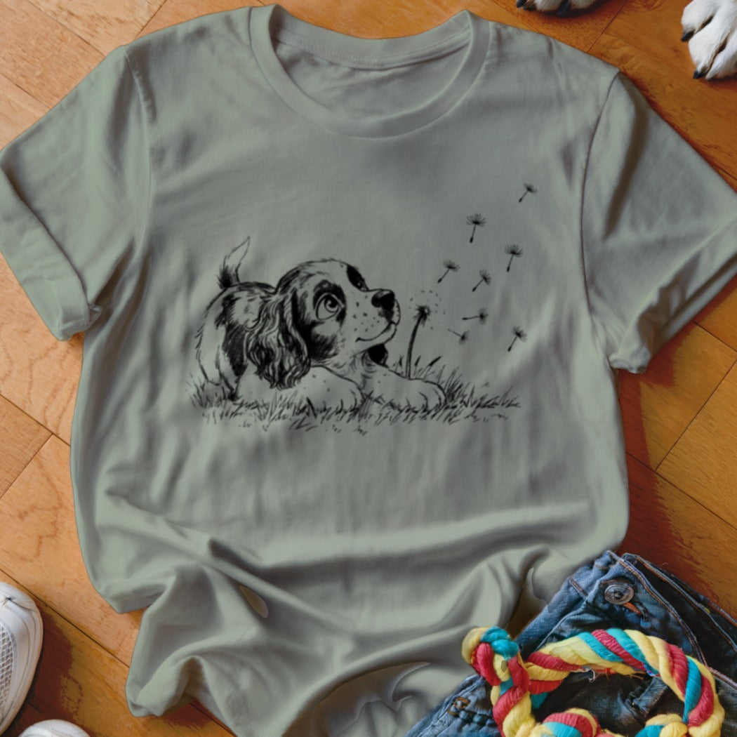Sage green crewneck t-shirt featuring a black line art graphic of a puppy watching dandelion seeds.