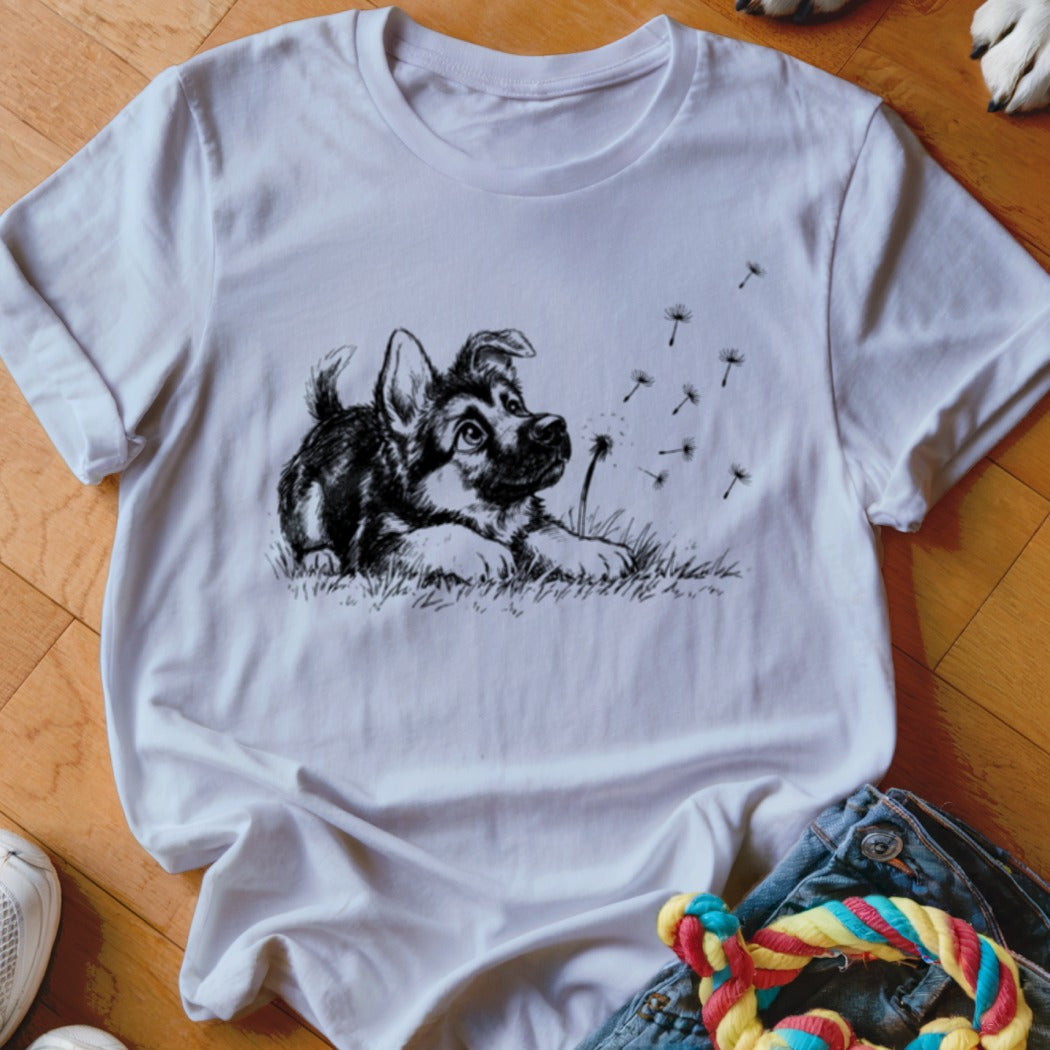 A white crew-neck t-shirt featuring a black graphic illustration of a German Shepherd puppy watching dandelion seeds blow away.