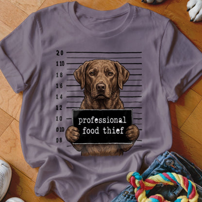 Brown dog mugshot with "professional food thief" sign on purple t-shirt.