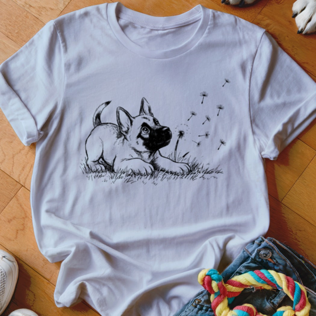White t-shirt featuring a black line drawing of a puppy playing with dandelions, laid flat on a wooden floor.