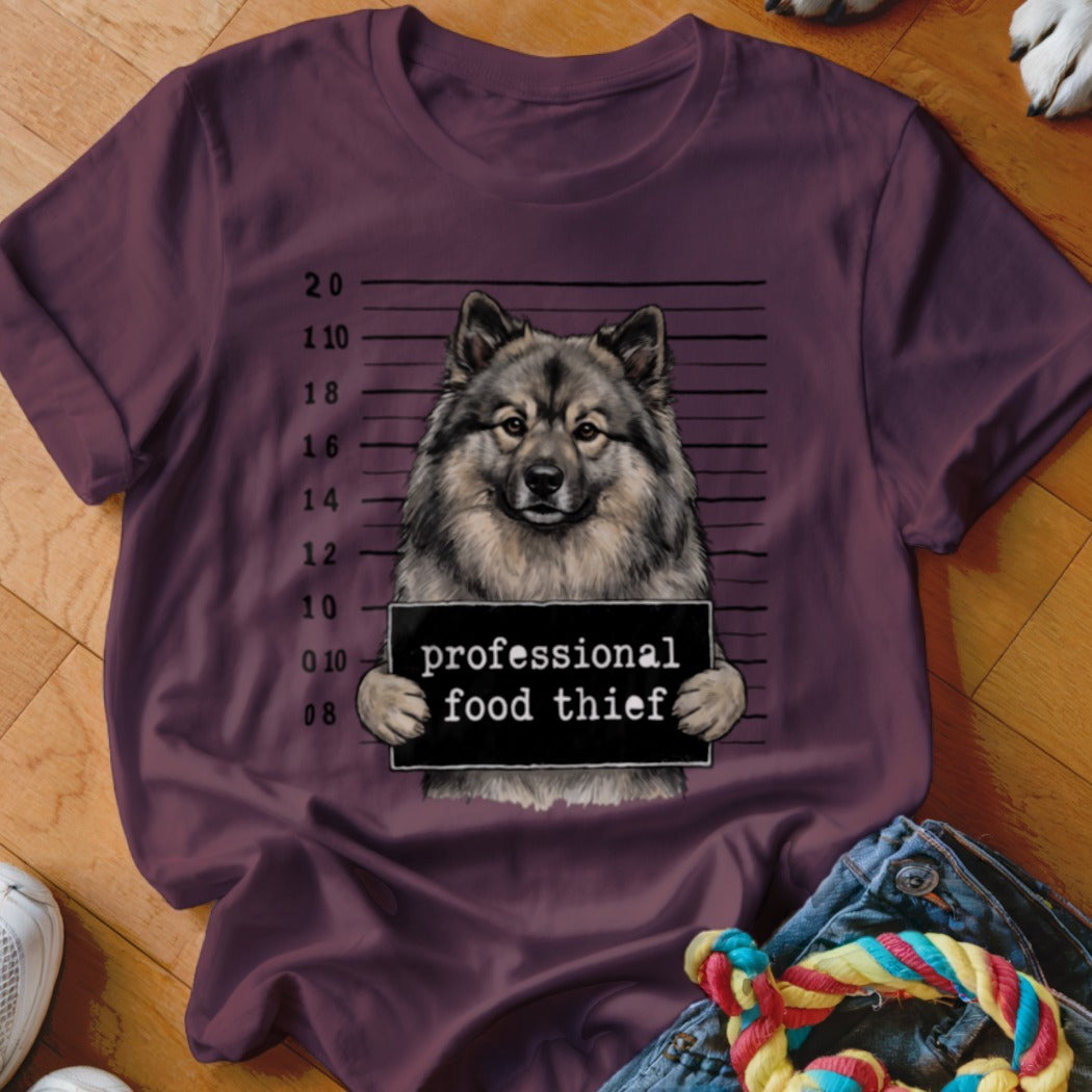 Keeshond dog mugshot holding a "professional food thief" sign on a maroon t-shirt.