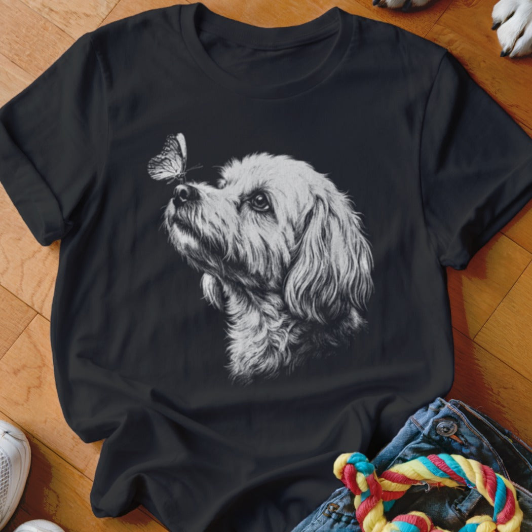 A black t-shirt features a white sketch of a fluffy dog looking at a butterfly on its nose.
