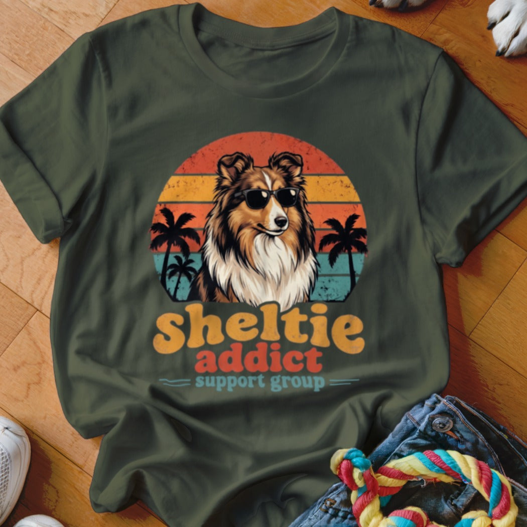 An olive green t-shirt featuring a retro sunset graphic with a Sheltie wearing sunglasses and palm trees.