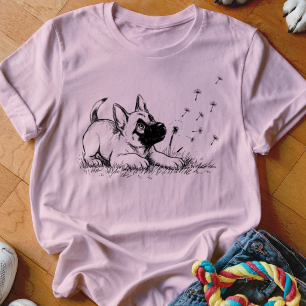 A pink crewneck t-shirt featuring a black line drawing of a puppy looking at floating dandelion seeds.
