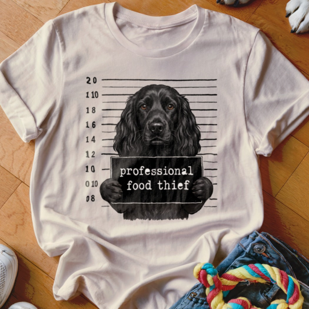 Black dog mugshot with "professional food thief" sign on a cream t-shirt.
