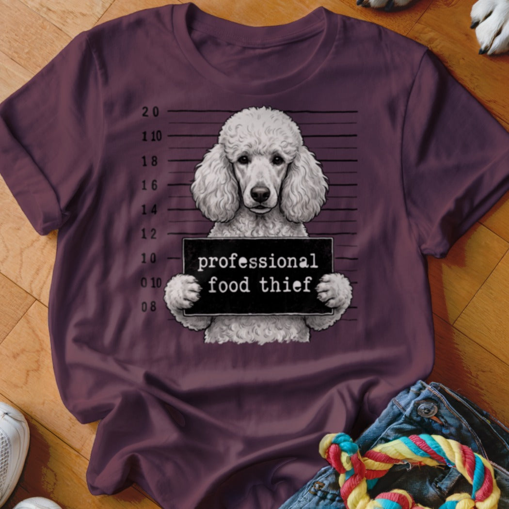 White poodle in mugshot holding "professional food thief" sign on maroon t-shirt.