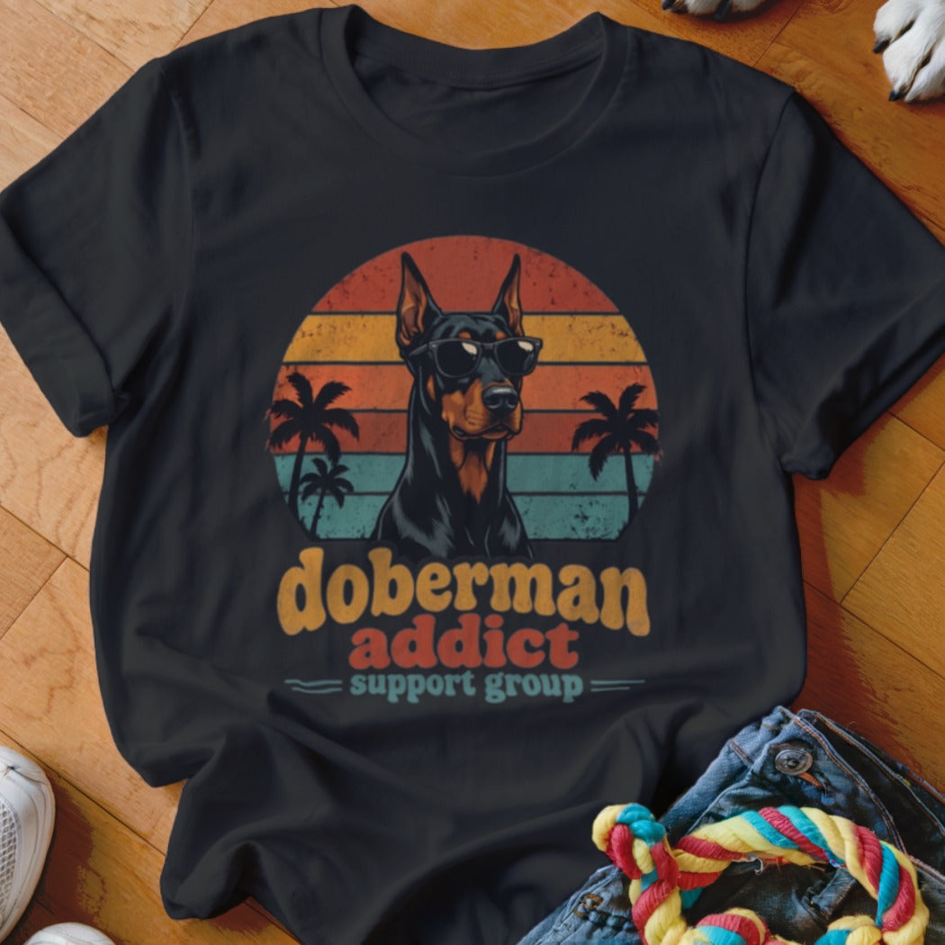 Black t-shirt featuring a retro Doberman graphic with sunglasses, palm trees, and sunset stripes, plus text reading doberman addict support group.