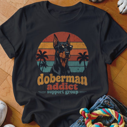Black t-shirt featuring a retro Doberman graphic with sunglasses, palm trees, and sunset stripes, plus text reading doberman addict support group.