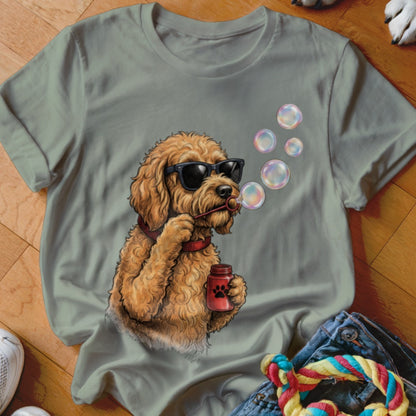 A sage green t-shirt featuring an illustration of a fluffy golden doodle wearing sunglasses and blowing bubbles.