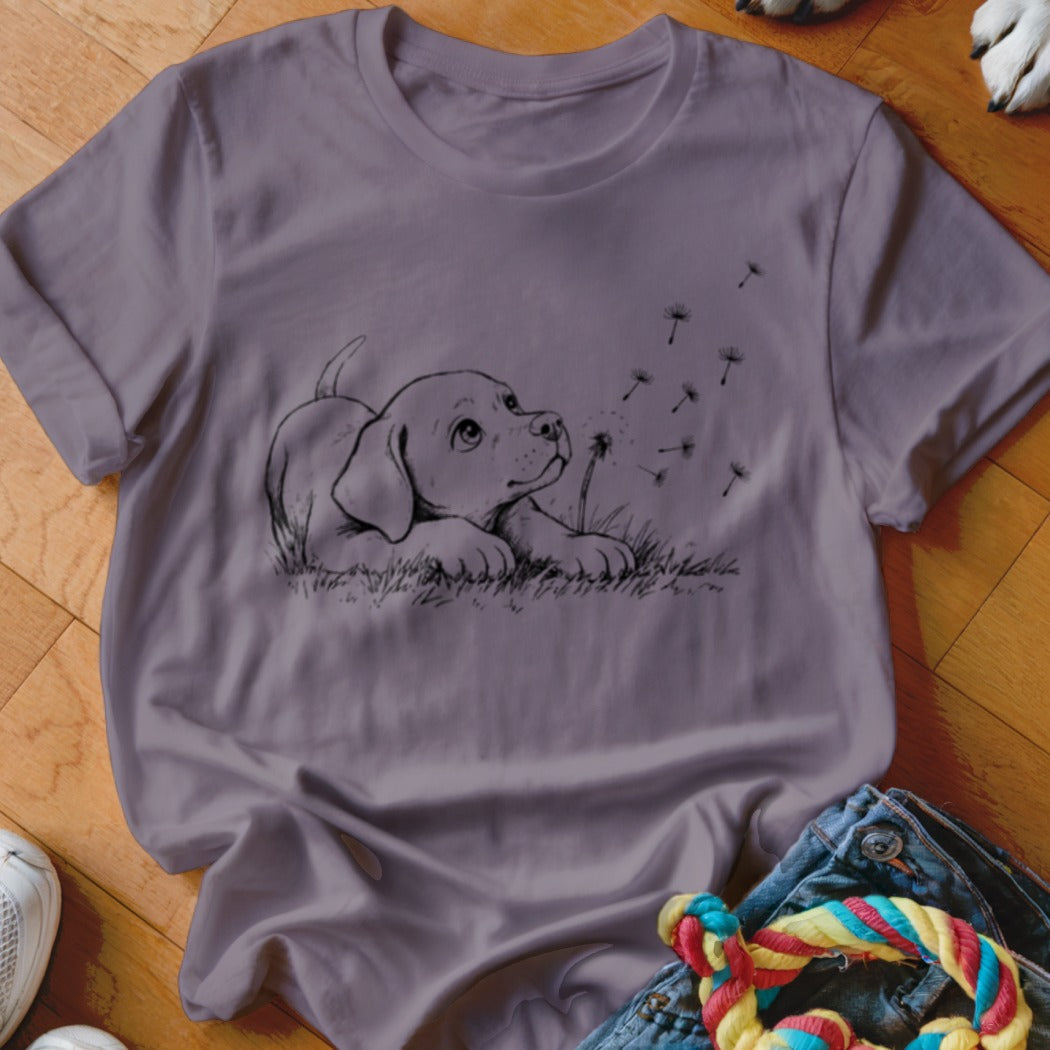 A purple short-sleeved t-shirt featuring a black line art graphic of a puppy watching floating dandelion seeds.