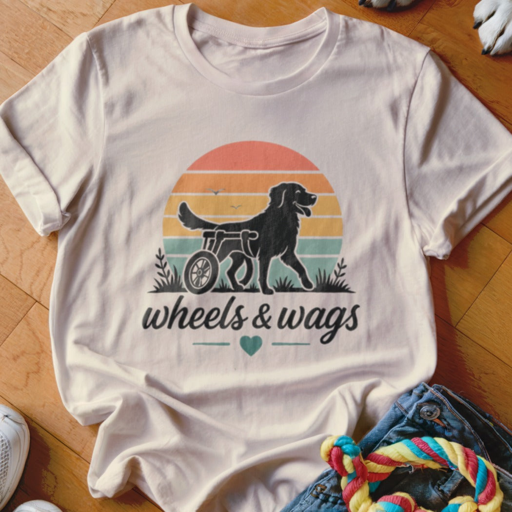 T-shirt front featuring a black dog in a wheelchair silhouette against a retro sunset with text "wheels & wags".