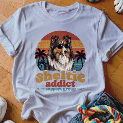 White t-shirt featuring a Sheltie wearing sunglasses in front of a sunset, with palm trees and text.