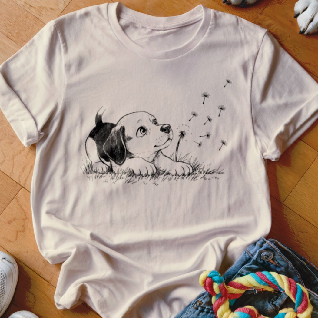 A light pink t-shirt features a black line sketch of a puppy looking at floating dandelion seeds.