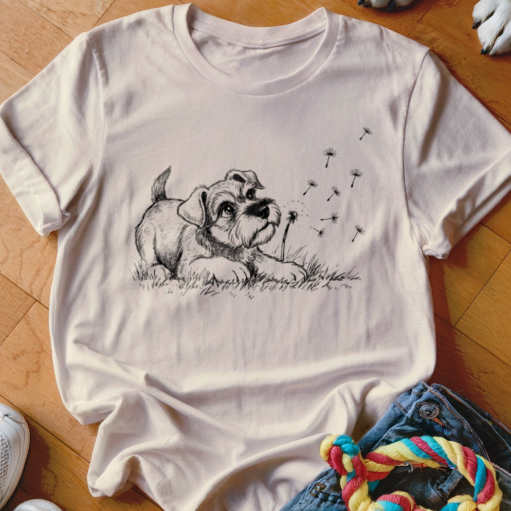 A light pink t-shirt featuring a black line drawing of a puppy watching blowing dandelion seeds.