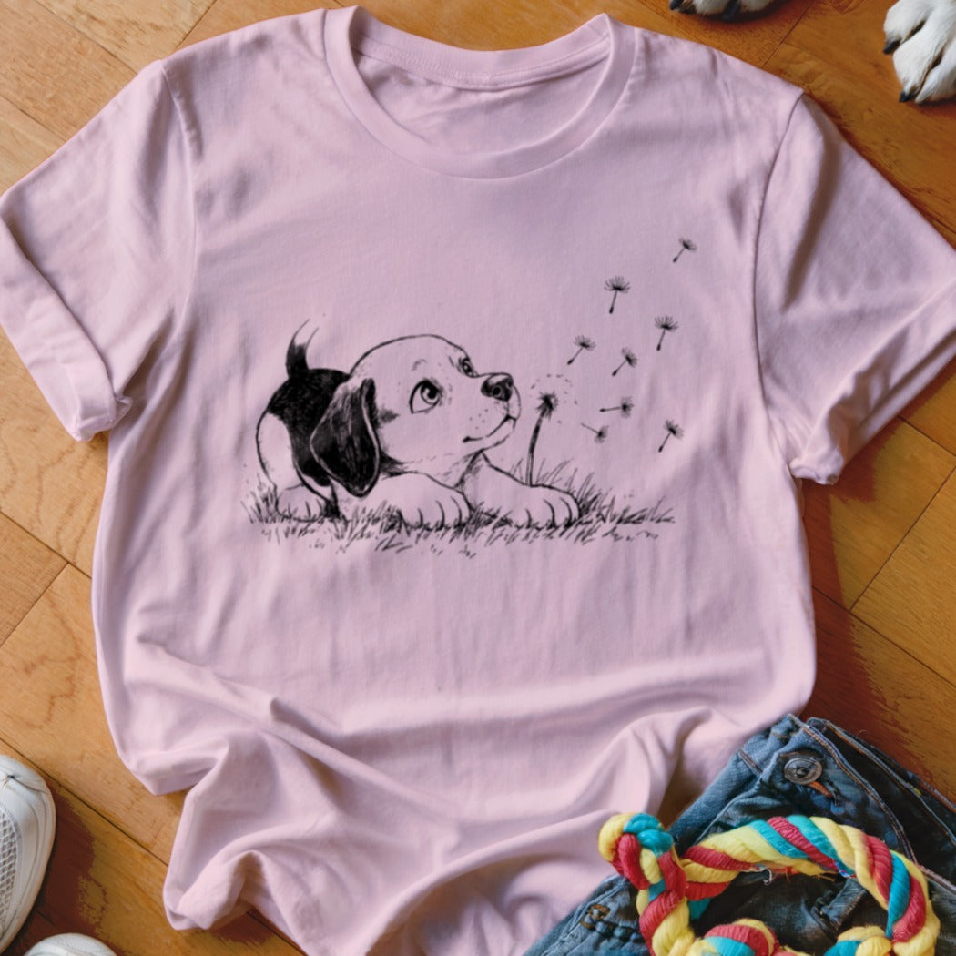A pink t-shirt featuring a line drawing of a puppy watching floating dandelion seeds.