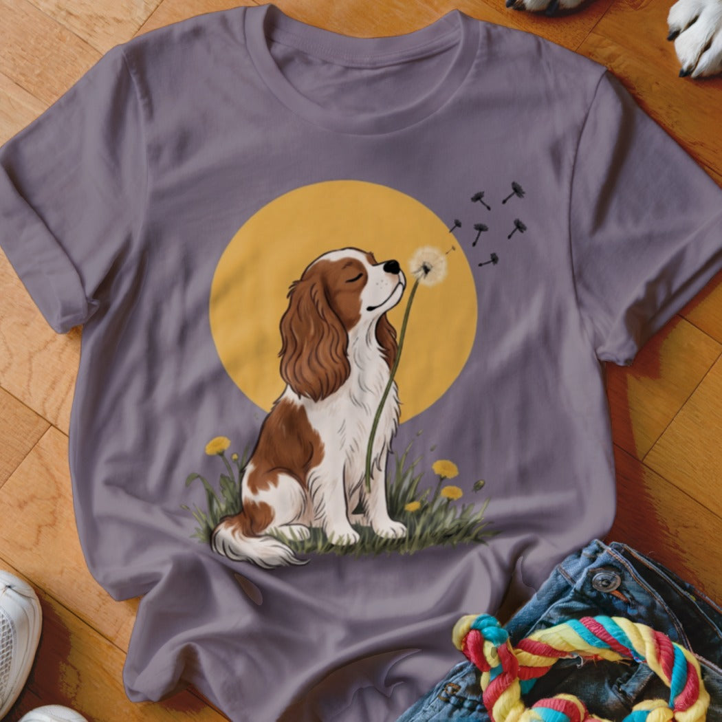 Purple t-shirt featuring an illustration of a Cavalier King Charles Spaniel blowing dandelion seeds in front of a yellow sun.