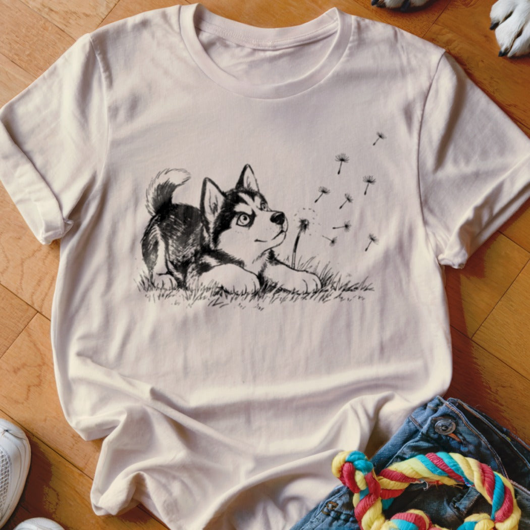A cream-colored t-shirt with a black line drawing of a husky puppy playing with dandelion seeds.