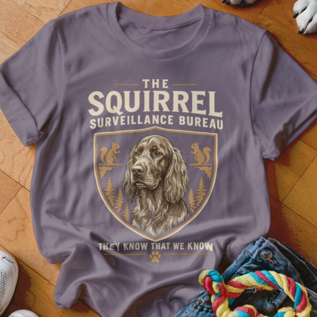 A purple t-shirt featuring a crest with a dog illustration and the text THE SQUIRREL SURVEILLANCE BUREAU.