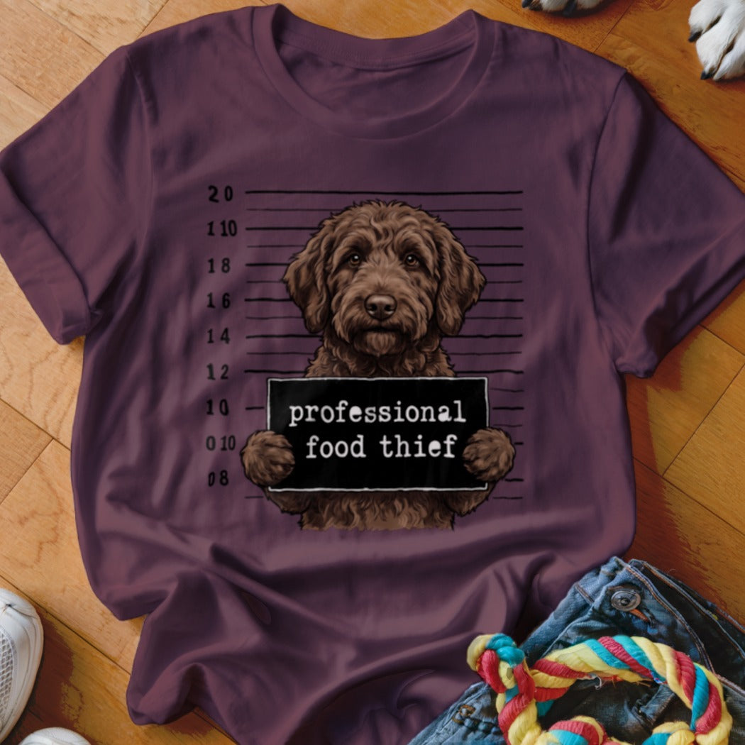 Brown dog in mugshot holding "professional food thief" sign on a maroon t-shirt.